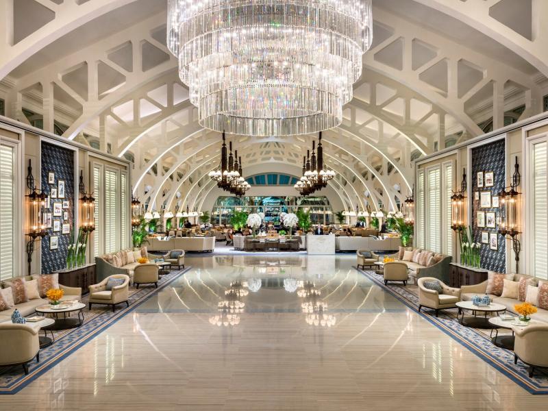 The Fullerton Bay Hotel grand arched lobby dazzles with a cascading crystal chandelier, polished marble floors, and elegant cream and gold seating framed by white vaulted beams.