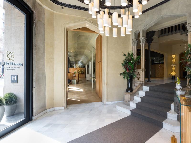 Monument Hotel grand, light-filled lobby showcases sculptural chandeliers, arched stone architecture, and polished marble floors in serene ivory and warm beige tones, accented by verdant greenery.