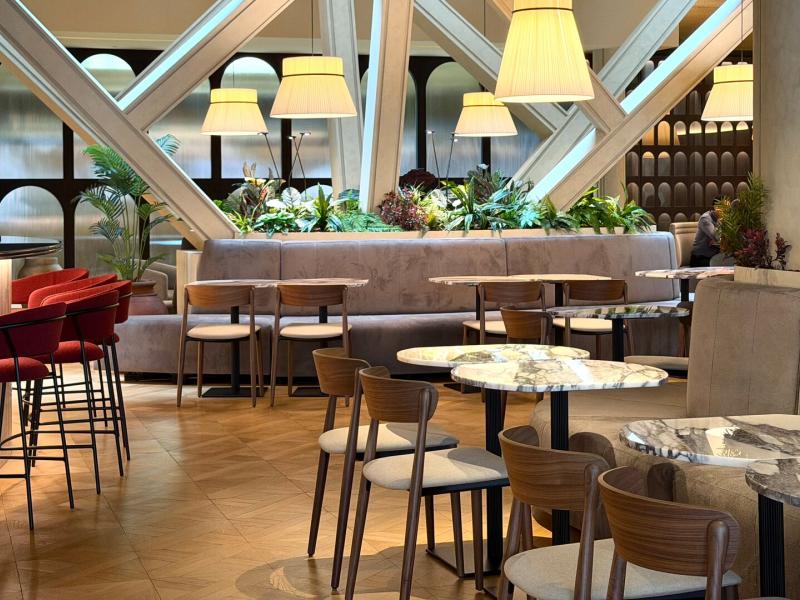 A chic, plant-filled lobby lounge pairs marble-topped tables and warm wood seating with soft taupe banquettes, elevated by glowing ivory pendant lights and rich terracotta accents.