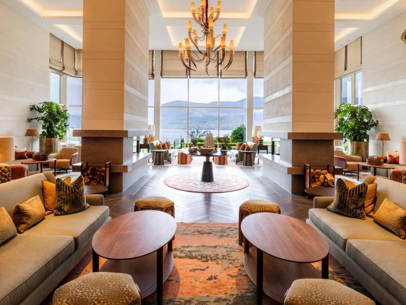 The Europe Hotel & Resort grand lobby adorned with soaring fireplaces, elegant chandeliers, and plush seating in soft beige, taupe, and warm copper tones, framed by picturesque lake and mountain views.