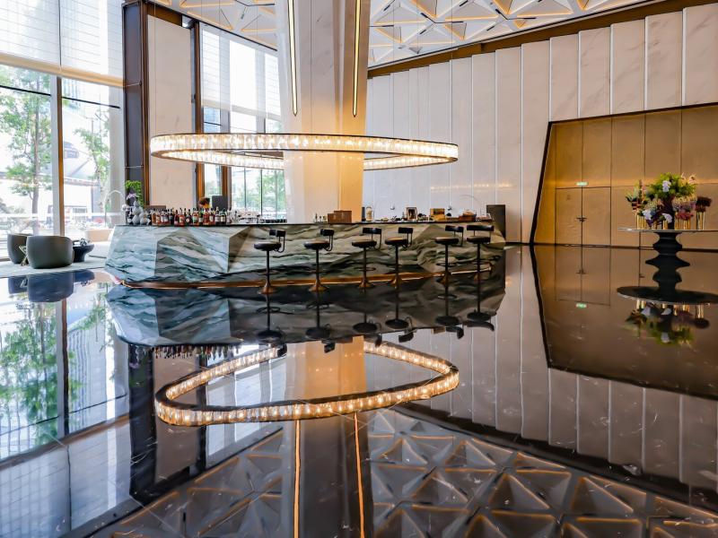 The lobby is paired with the Café Bar Lounge with a sleek marble-clad bar, dramatic statement chandelier, and glossy black reflective floors blend seamlessly with warm gold and crisp white hues.