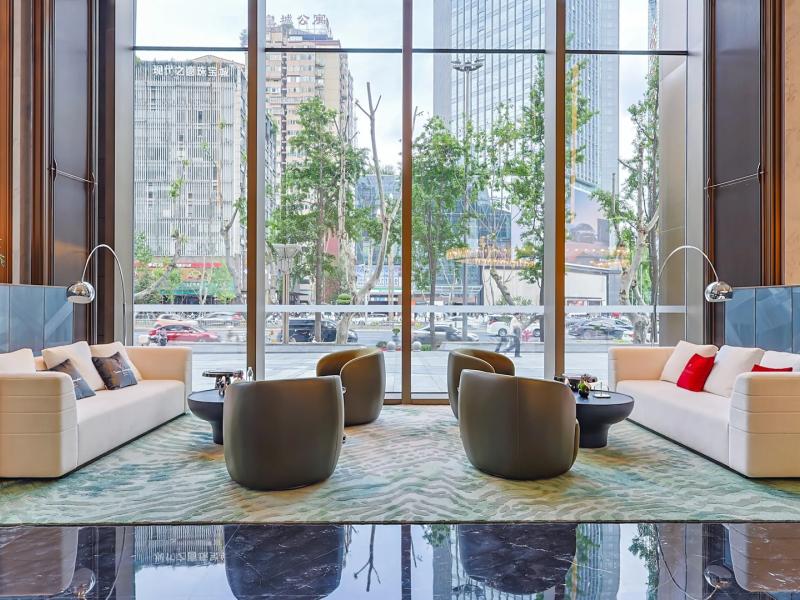 Tonino Lamborghini Hotel Chengdu lobby presents floor-to-ceiling windows, plush ivory sofas, and contemporary charcoal lounge chairs, accented by soft sage green and vibrant ruby-red touches.