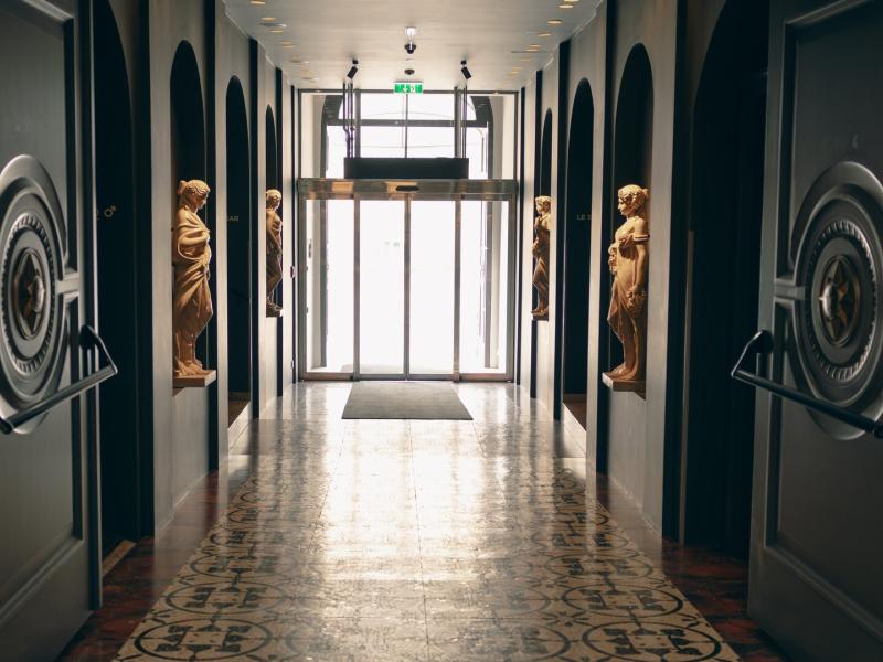 Another grand corridor showcases stately sculptures, dramatic archways, and intricate patterned flooring highlighted by deep charcoal and warm gold accents.