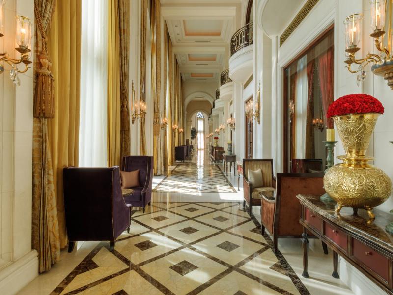 A grand corridor inside The Leela Palace New Delhi adorned with soaring gold drapery, gleaming marble floors, and ornate crystal sconces, accented by rich red florals and elegant emerald-green details.