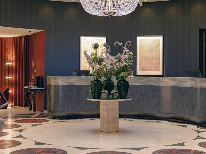 Hotel Sans Souci Wien elegant lobby welcomes guests with a sparkling crystal chandelier, contemporary floral arrangements, and refined charcoal and silver tones accented by soft lavender hues.