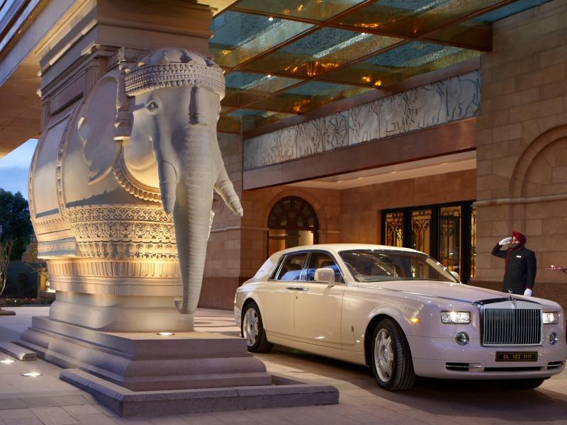 The Leela Palace New Delhi grandeur entrance with real life-size sculpted ivory elephant statues and a gleaming glass porte-cochère, accented by warm gold lighting and rich crimson details.