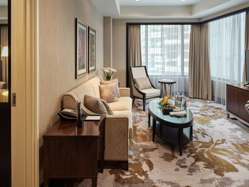 An elegant One-Bedroom Suite living room blends creamy beige seating, rich walnut finishes, gold-accented carpeting, a glass coffee table, and expansive corner windows filled with natural light, all adjacent to a luxurious bedroom.