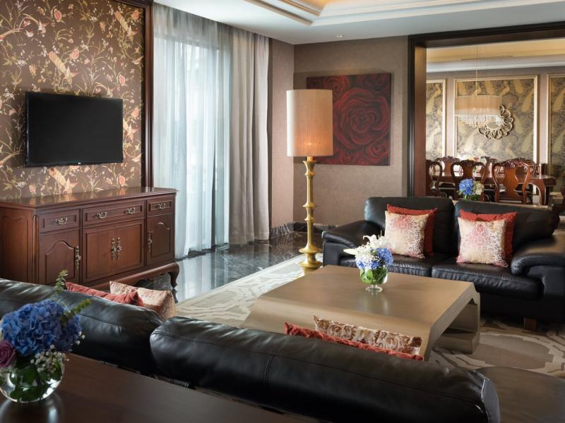 An opulent AYANA Suite living space pairs rich mahogany furnishings and black leather seating with floral artistry, crimson accents, blue hydrangeas, soft gold lighting, and an elegant adjoining dining room.