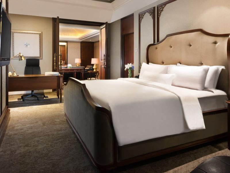 The AYANA Suite’s serene luxury bedroom blends crisp white linens and a dramatic dark wood bed with champagne-beige tones, plush seating, a sleek workspace, warm golden lighting, and softly filtered natural light.