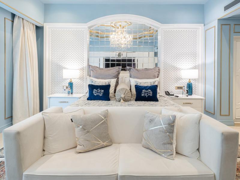 The Claridges Suite bedroom presents gilded powder-blue walls that frame a sumptuous bed with navy velvet accents, mirrored panels, crystal lighting, luminous white furnishings, and an elegant lounge sofa.