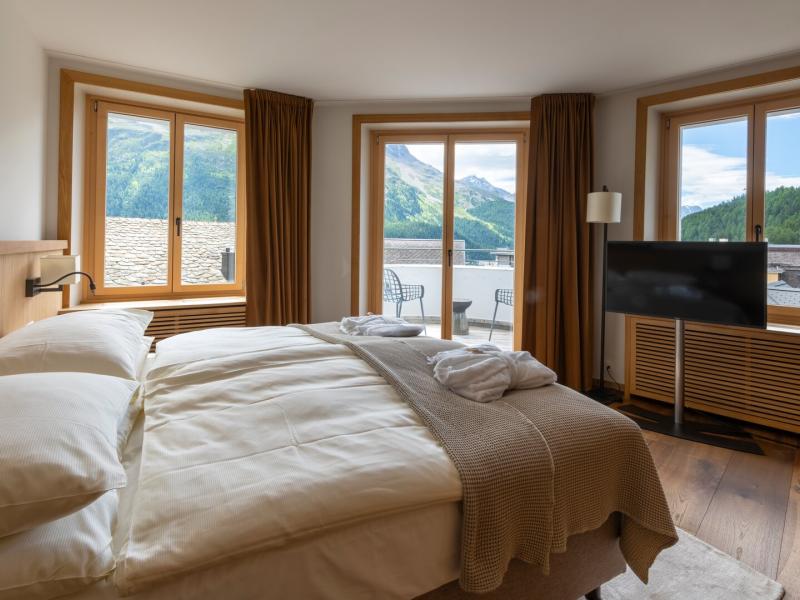 This Deluxe Suite offers ivory bedding, taupe textures, caramel drapes, colorful artwork, and a furnished balcony overlooking emerald mountains.