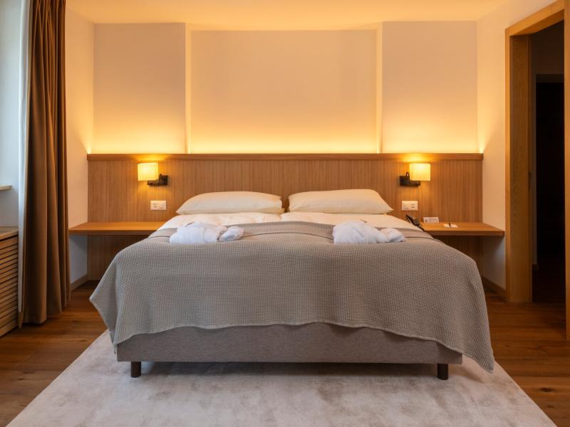 Golden backlit paneling and brass-toned sconces bathe a serene oak-framed bed in amber light in a Deluxe Suite, dressed in dove-gray knit, crisp white pillows, and waiting robes above a plush ivory rug.