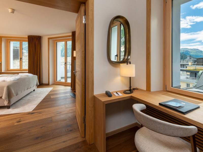 A Deluxe Suite features an oak writing desk with a bronze oval mirror and sandstone-toned chair overlooking sky-blue mountain vistas, while an open door reveals a serene bedroom of taupe linens and gleaming hardwood floors.
