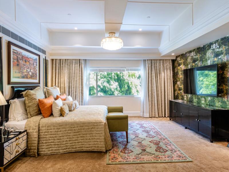 Champagne bedding, burnt-orange pillows, verdant mural walls, ebony furnishings, a crystal chandelier, garden views, and a richly patterned rug elevate this lavish bedroom of the Presidential Suite.