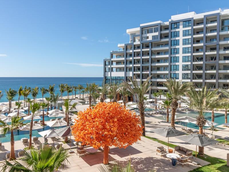 Garza Blanca Resort & Spa Los Cabos exterior featuring a striking orange tree centerpiece surrounded by palm-lined pools and sun loungers, with a sleek multi-story building overlooking the ocean.