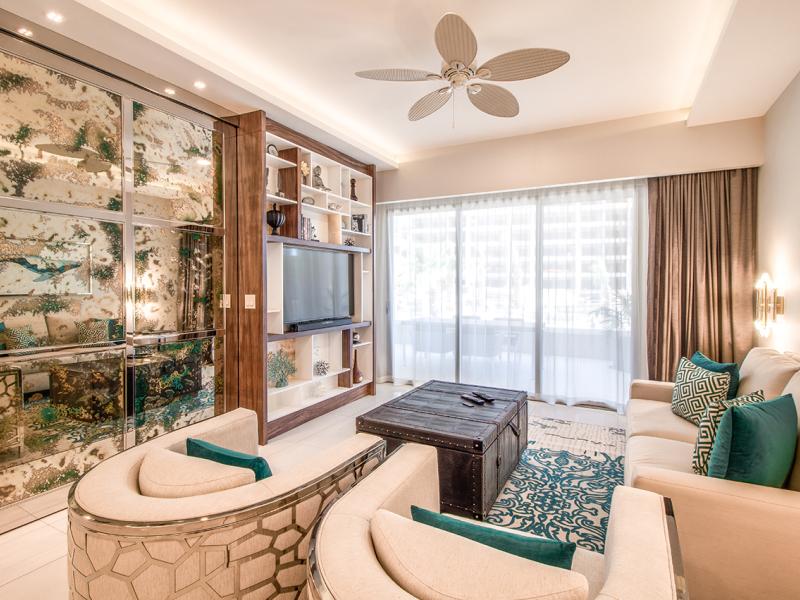 One Bedroom Suite chic, contemporary living area features cream seating with teal accents, a mirrored wall with nature-inspired design, and floor-to-ceiling windows flooding the space with natural light.