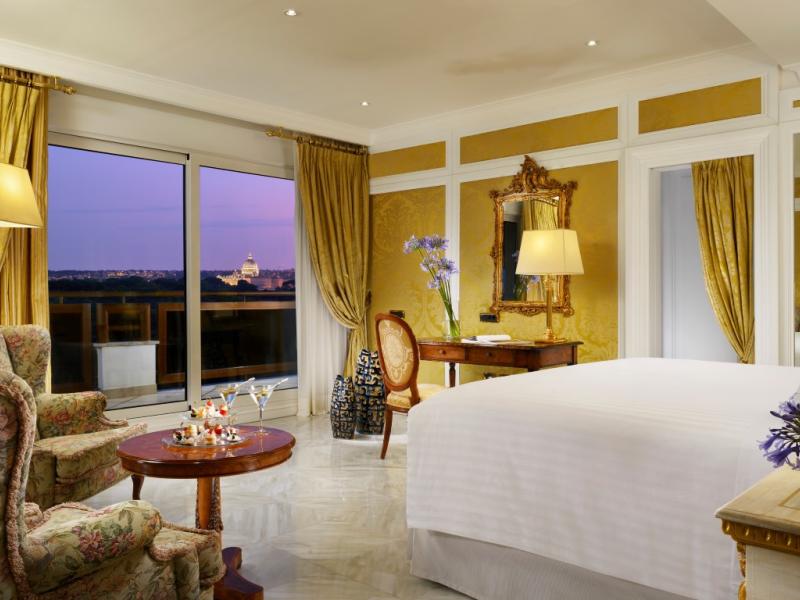 Parco dei Principi Grand Hotel & Spa’s Royal Suite bedroom features floor-to-ceiling windows, a cozy seating area, and elegant drapery, accented in soft gold, cream, and warm wood tones.