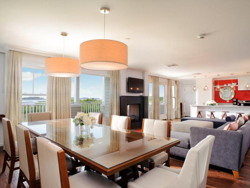 A Beach Suite expansive, sunlit living and dining space features a gleaming glass-top table, plush seating, and a fireplace backdrop, elevated by creamy ivory, slate gray, and coral accents.