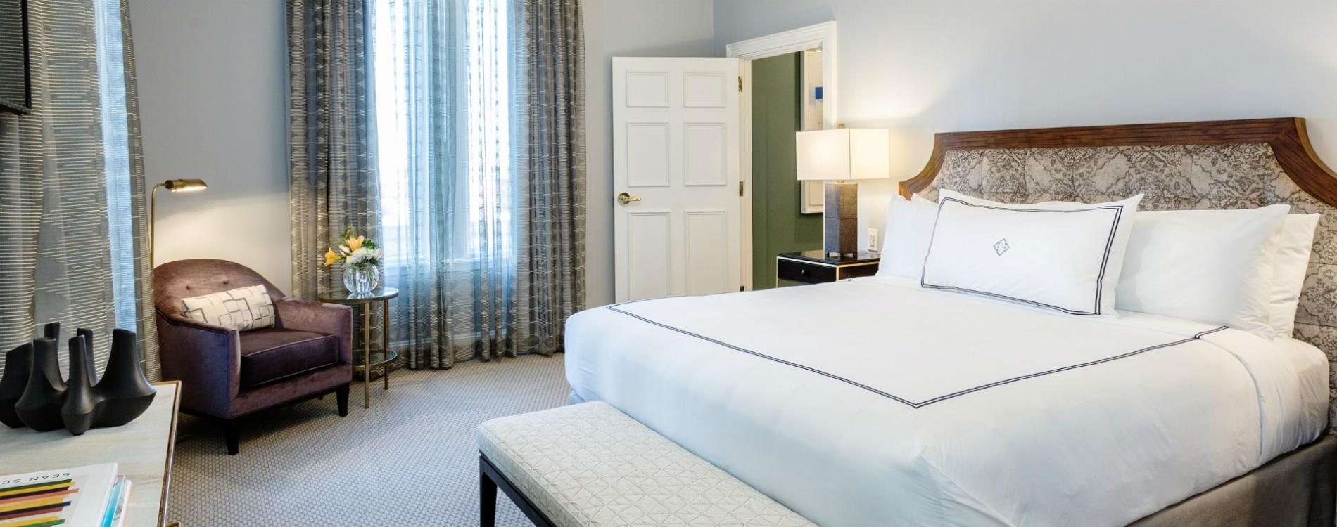 A hotel room with a large bed, comfortable chair, and window with curtains.